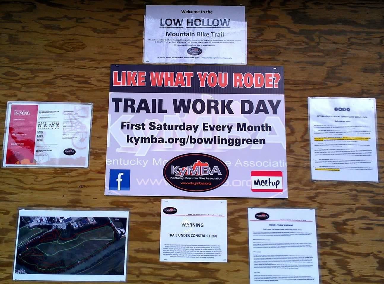 A bulletin board at the Low Hollow Mountain Bike Trail featuring various announcements and information. Highlights include a sign for Trail Work Day scheduled for the first Saturday of every month, a map of the trail layout, a warning about trail construction, and a welcome message from the Kentucky Mountain Bike Association (KyMBA). Additional details include social media links and instructions for volunteers. Low Hollow mountain bike trail.