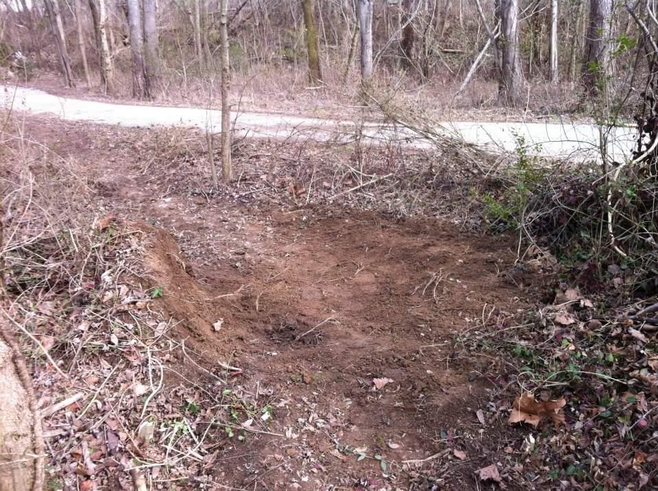 A cleared patch of dirt surrounded by sparse vegetation and trees, with a dirt pathway visible in the background. The area appears to be disturbed, with some leaves scattered around. Low Hollow mountain bike trail.