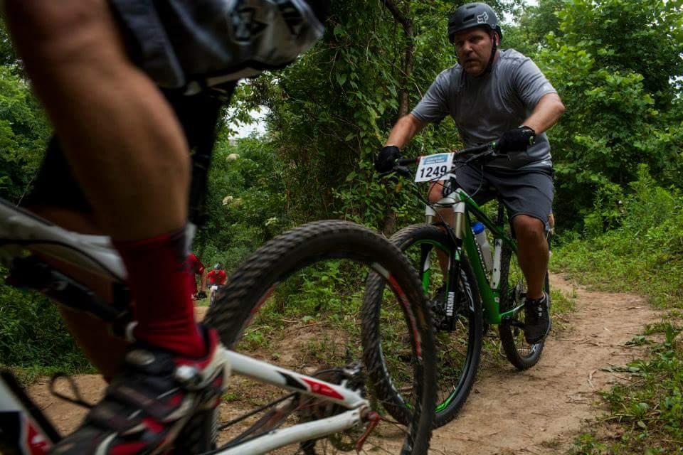 A man in a gray shirt and shorts rides a mountain bike along a dirt trail surrounded by greenery. Another cyclist's bike partially enters the frame in the foreground, showing detail of the wheel and leg in motion. The scene captures an outdoor cycling event or competition. Low Hollow mountain bike trail.