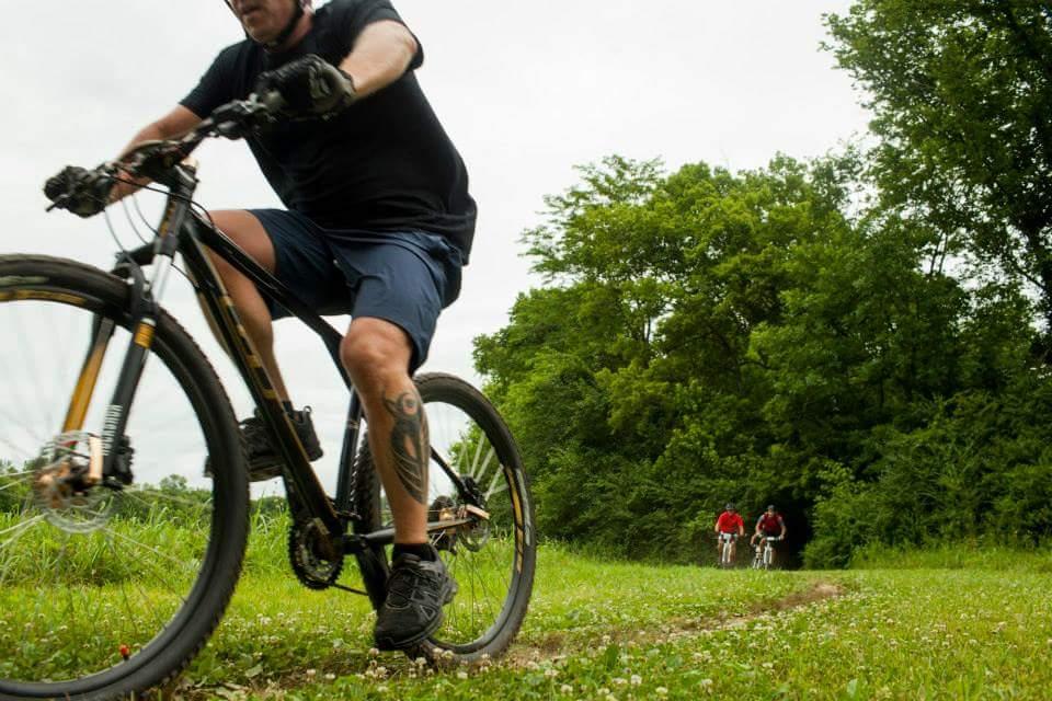 A man riding a mountain bike on a grassy trail, with two people on bikes in the background. The scene is surrounded by lush greenery under a cloudy sky, suggesting an outdoor cycling activity. Low Hollow mountain bike trail.