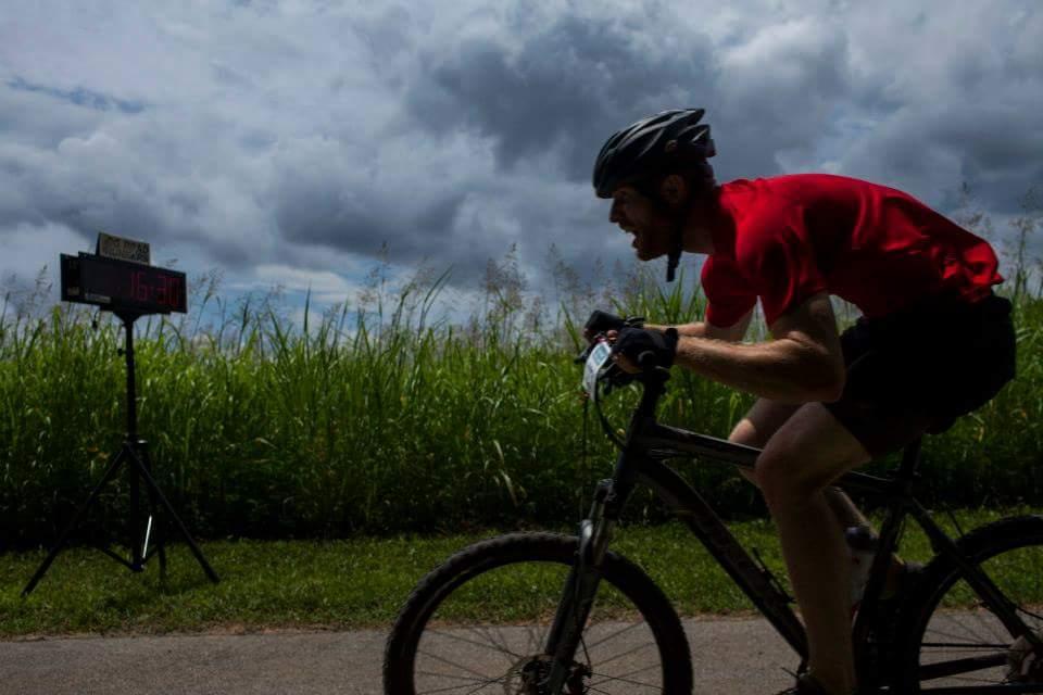 A cyclist wearing a red shirt and a helmet rides a mountain bike along a path, with tall grass on either side. A digital timer displaying "16:30" is visible in the background under a cloudy sky. Low Hollow mountain bike trail.