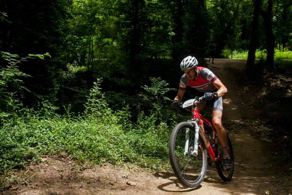 A mountain biker navigating a dirt trail through a lush, green forest. The cyclist is wearing a helmet and cycling attire, focused on the path ahead as they ride a red mountain bike. Sunlight filters through the trees, creating a vibrant, natural atmosphere. Low Hollow mountain bike trail.