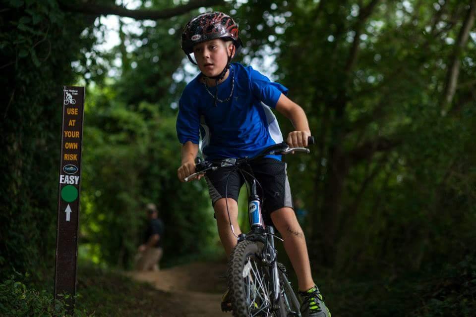 A young boy in a blue shirt rides a mountain bike along a dirt trail surrounded by lush greenery. A trail sign beside him indicates the route is easy and warns users to proceed at their own risk. In the background, another person can be seen walking on the trail. Low Hollow mountain bike trail.