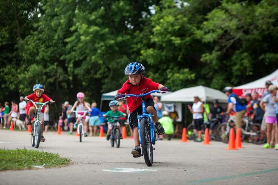 Children participating in a bike racing event, with several riders in action on a paved path. In the foreground, a boy in a red shirt and blue helmet is pedaling hard, while other young cyclists ride in the background. A crowd of spectators, including adults and kids, watch from the sidelines, surrounded by orange cones marking the course. Lush greenery is visible in the background. Low Hollow mountain bike trail.