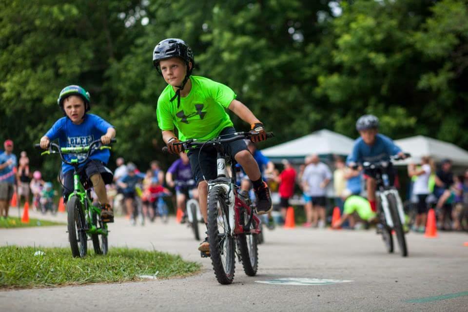 Children participating in a biking competition on a paved path, with orange cones marking the course. One boy in a bright green shirt rides confidently in the foreground, while another boy in a blue shirt follows closely behind. In the background, other participants and spectators can be seen amidst trees and tents, creating a lively atmosphere. Low Hollow mountain bike trail.