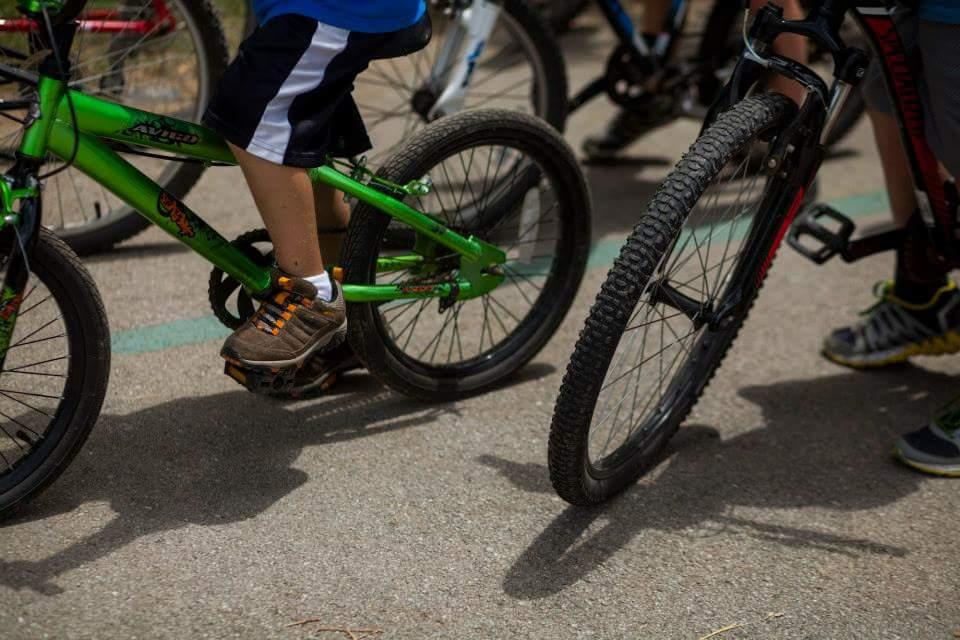 A close-up view of children's legs riding bicycles, featuring a green bike and a black bike. The scene captures the motion of biking on a paved surface, with focus on the footwear and pedals of the riders. Low Hollow mountain bike trail.
