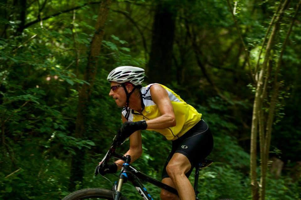 A cyclist in a yellow and white jersey rides a mountain bike through a lush, green forest trail, showcasing determination and focus while navigating the terrain. Low Hollow mountain bike trail.