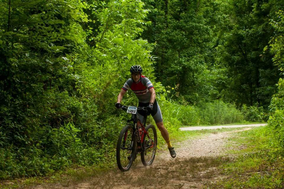 A cyclist in a red and gray jersey rides on a dirt path surrounded by greenery, kicking up dust as they navigate through the forested area. Low Hollow mountain bike trail.