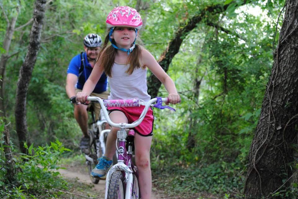 A young girl wearing a pink helmet rides a bicycle along a wooded trail, smiling as she navigates the path. A man in a blue shirt is biking behind her, both surrounded by lush green foliage. Low Hollow mountain bike trail.