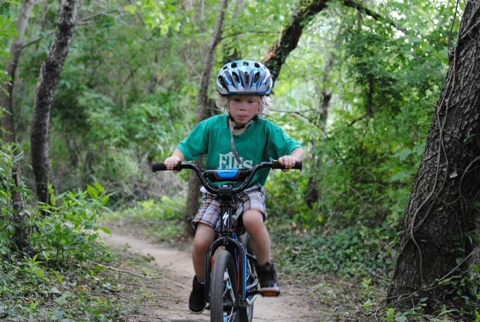 A young child riding a bicycle on a dirt path surrounded by lush greenery. The child is wearing a blue helmet and a green shirt with shorts, appearing focused while cycling. Low Hollow mountain bike trail.