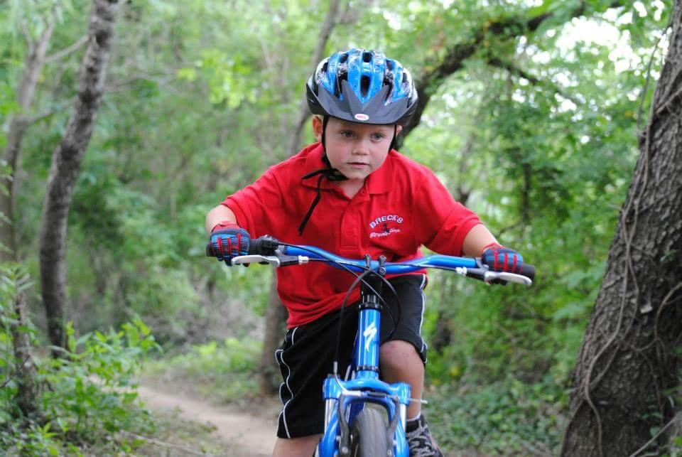 A young boy in a red shirt and black shorts rides a blue bicycle on a dirt path surrounded by greenery. He wears a black bicycle helmet and gloves, focusing intently as he navigates the trail. Low Hollow mountain bike trail.