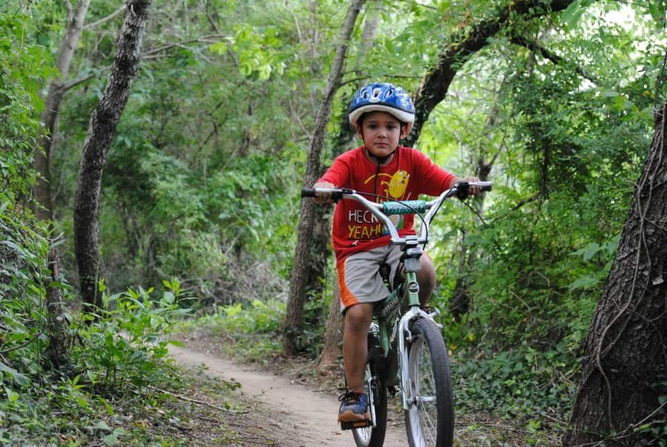 A young boy wearing a blue helmet and a red t-shirt is riding a green bicycle on a dirt trail surrounded by lush greenery and trees. Low Hollow mountain bike trail.