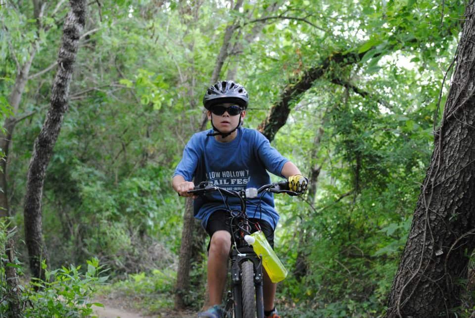 A young boy wearing sunglasses and a helmet rides a mountain bike on a dirt trail surrounded by green trees and lush foliage. He is focused on the path ahead, showcasing a sense of adventure in an outdoor setting. Low Hollow mountain bike trail.