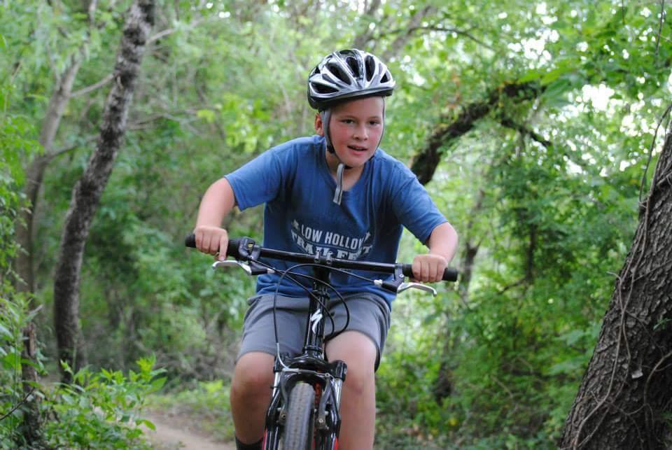 A young boy riding a bicycle on a dirt trail surrounded by lush green trees. He is wearing a black helmet and a blue t-shirt, looking focused and enjoying the ride. Low Hollow mountain bike trail.