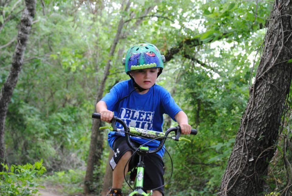 A young boy wearing a colorful helmet rides a green bicycle on a dirt path surrounded by trees and greenery. He appears focused as he navigates the trail, dressed in a blue shirt with a bold white graphic. Low Hollow mountain bike trail.
