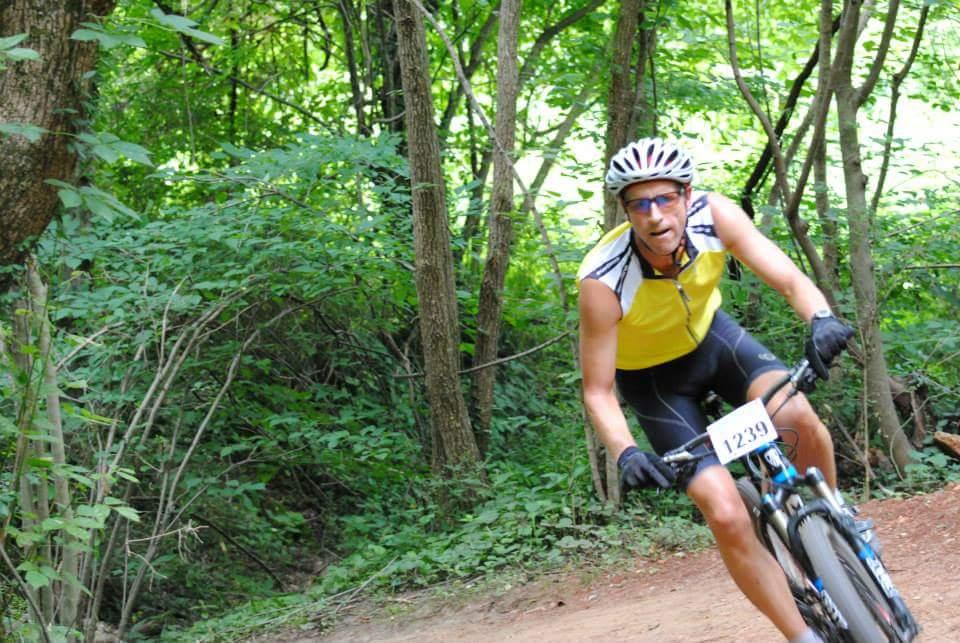 A person riding a mountain bike along a dirt trail in a lush, green forest. The cyclist is wearing a yellow and black jersey and a helmet, with a visible race number on their bike. The scene captures the activity in a vibrant outdoor setting. Low Hollow mountain bike trail.