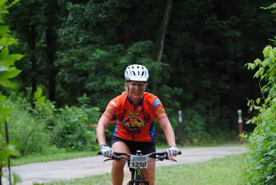 A woman wearing a bright orange cycling jersey and a white helmet rides her mountain bike on a wooded path, surrounded by lush greenery. She appears focused and determined as she pedals along the trail. Low Hollow mountain bike trail.