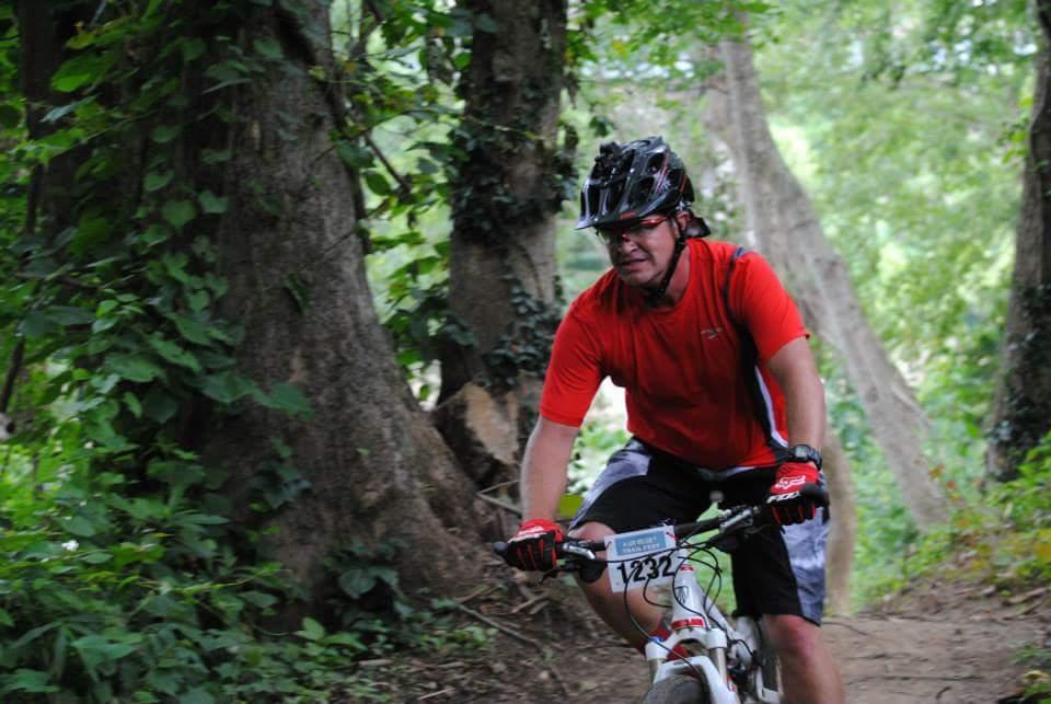 A person riding a mountain bike on a dirt trail surrounded by trees and greenery. They are wearing a red shirt, black shorts, and a helmet, with a race number visible on the bike. Low Hollow mountain bike trail.