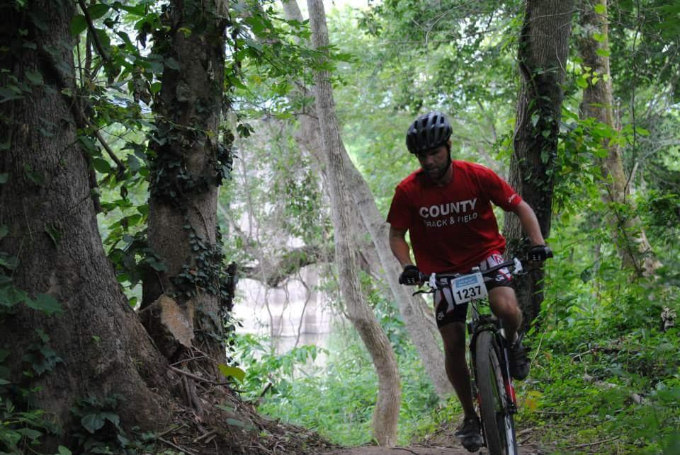 A person riding a mountain bike on a wooded trail, wearing a red shirt with "COUNTY TRACK & FIELD" printed on it and a helmet. The background features tall trees and lush greenery, creating a natural setting. Low Hollow mountain bike trail.