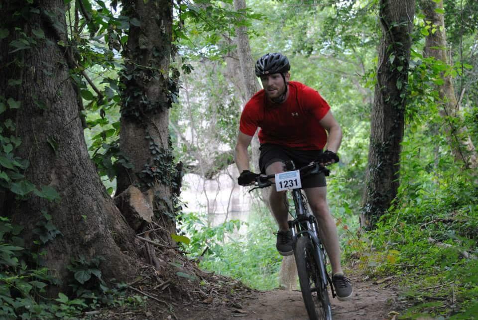 A cyclist in a red shirt and black shorts rides a mountain bike on a narrow dirt trail surrounded by lush green trees and foliage. The biker is wearing a helmet and gloves, with a race number displayed on the front of the bike. Low Hollow mountain bike trail.