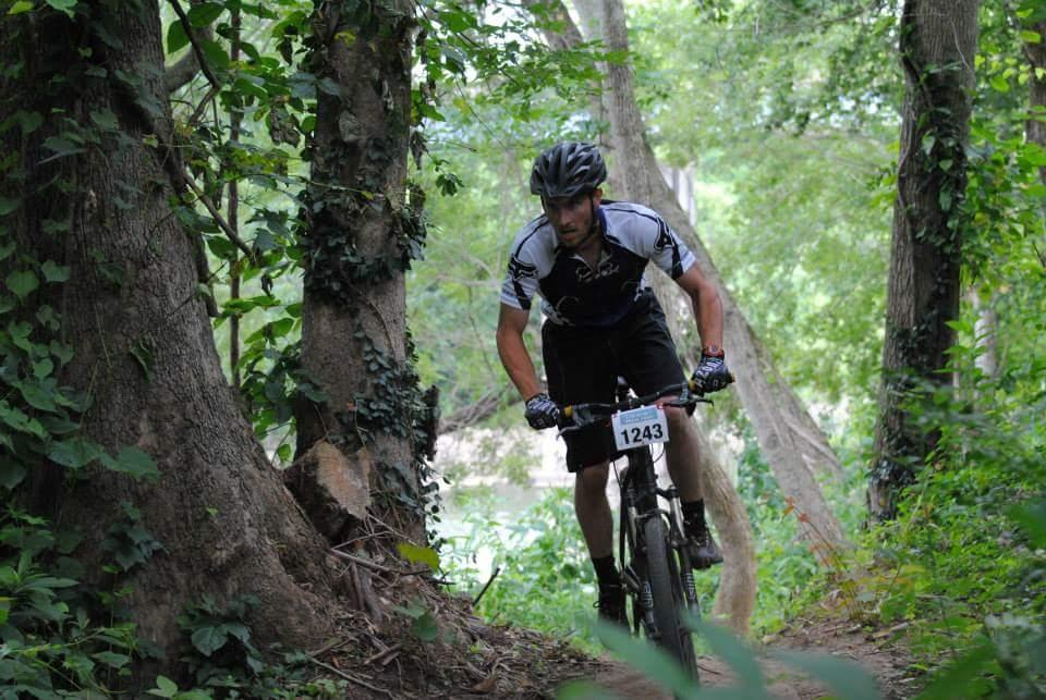 A mountain biker navigating a wooded trail, surrounded by lush greenery and trees. The cyclist is wearing a black and white jersey with a number 1243 displayed on the front. The scene captures the dynamic action of biking through a natural environment. Low Hollow mountain bike trail.