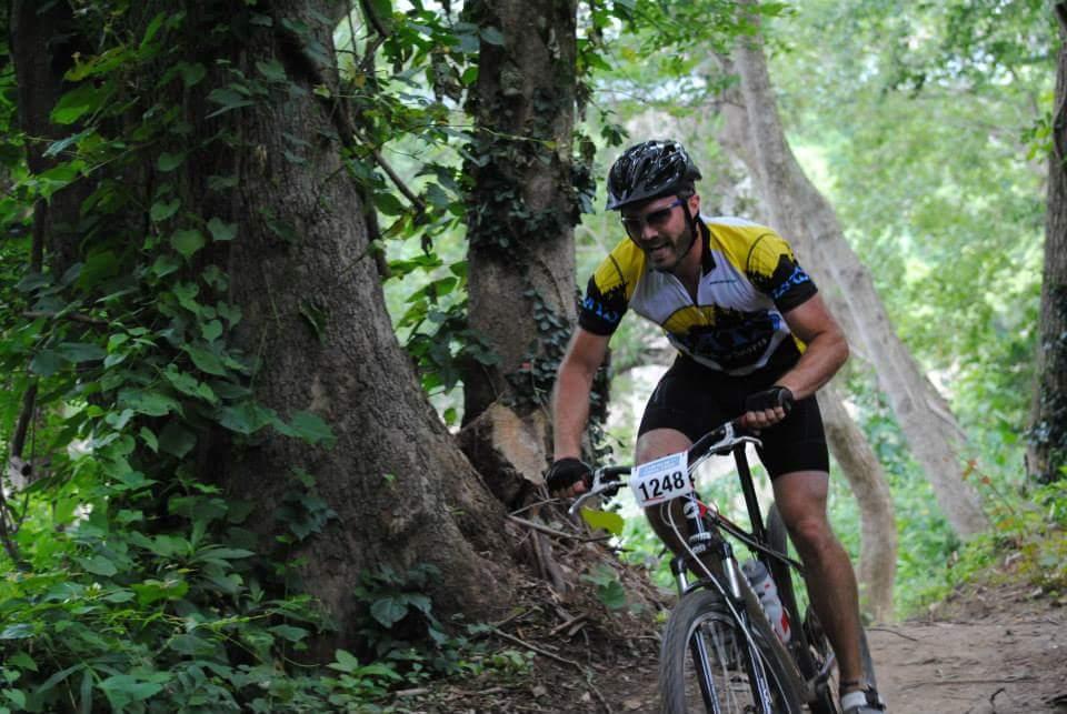 A mountain biker in a colorful jersey riding a bike along a dirt trail surrounded by lush greenery and trees. The cyclist appears focused and is climbing a steep section of the path, with a race number displayed on the bike. Low Hollow mountain bike trail.
