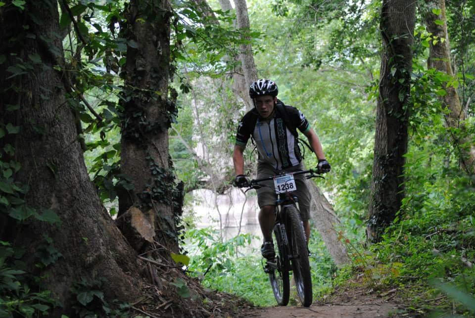 A person riding a mountain bike on a dirt trail surrounded by trees and greenery. The rider is wearing a helmet and sports attire, with a race number visible on their jersey. Low Hollow mountain bike trail.