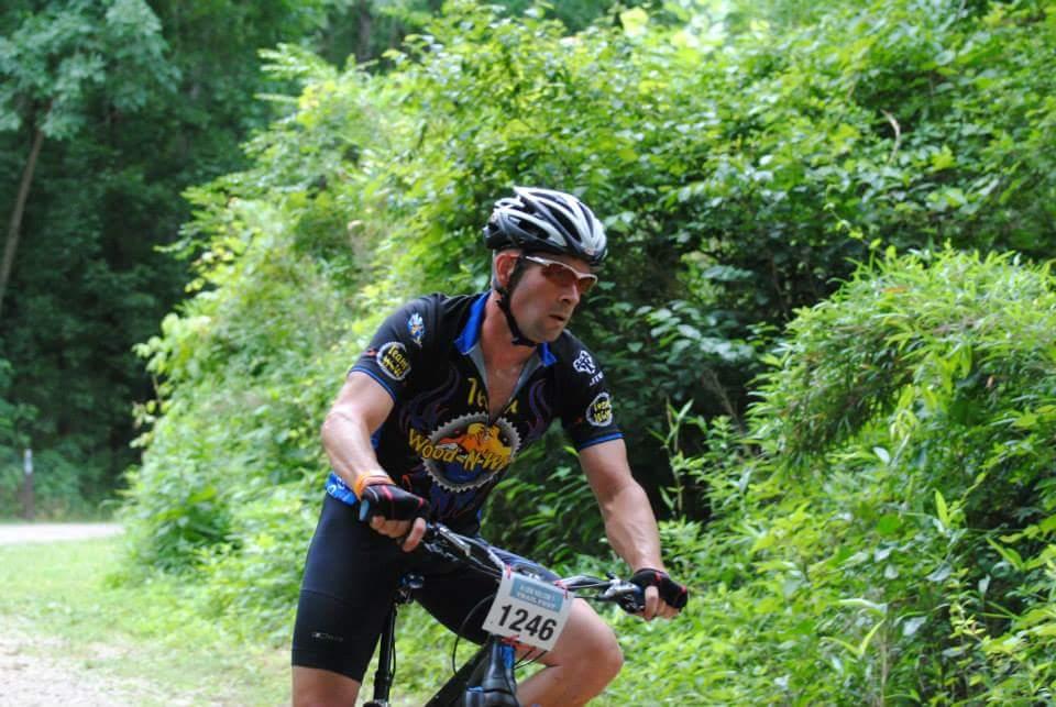 A cyclist wearing a black and blue jersey with the number 1246 rides on a dirt path surrounded by lush green foliage. The cyclist is focused and appears to be navigating through a wooded area. Low Hollow mountain bike trail.
