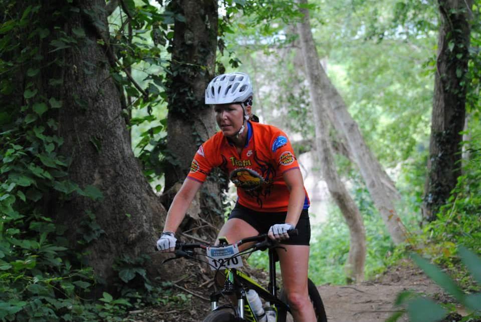 A female mountain biker in an orange jersey and white helmet rides a bike along a wooded trail, surrounded by lush greenery and trees. She appears focused and determined as she navigates the path in a natural outdoor setting. Low Hollow mountain bike trail.
