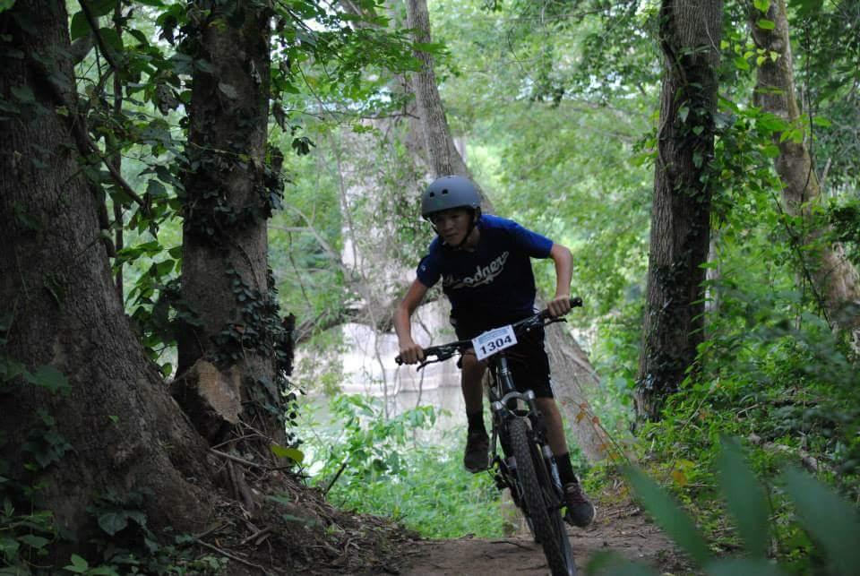 A young cyclist in a blue shirt and helmet rides a mountain bike down a wooded trail, surrounded by lush green trees and plants. The bike's number plate, 1304, is visible as the rider navigates the path. Low Hollow mountain bike trail.