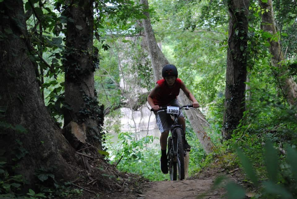 A young boy riding a mountain bike on a dirt trail surrounded by trees and greenery. He is wearing a red shirt, shorts, and a helmet, and has a race number displayed on his shirt. The scene captures the excitement of biking in a natural setting. Low Hollow mountain bike trail.
