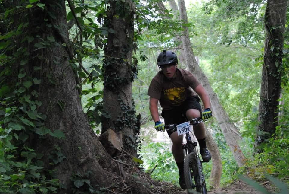 A mountain biker navigating a dirt trail surrounded by lush greenery and trees. The rider wears a helmet and protective gear, showcasing a focused expression as they pedal downhill. The scene captures the excitement of mountain biking in a natural environment. Low Hollow mountain bike trail.