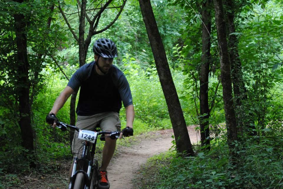A person riding a mountain bike on a dirt trail surrounded by lush green foliage and trees. The cyclist is wearing a helmet and a sporty outfit, focused on navigating the path. A numbered tag is visible on the bike. Low Hollow mountain bike trail.