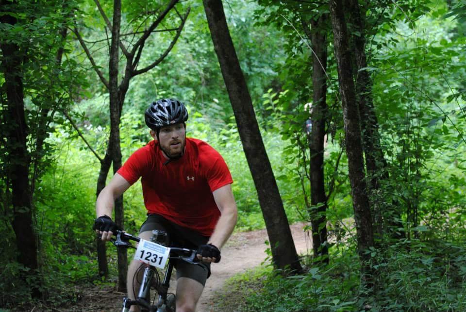 A mountain biker wearing a red shirt and helmet rides along a dirt trail surrounded by lush green vegetation and trees. The biker is focused, and a race number is visible on the front. Low Hollow mountain bike trail.