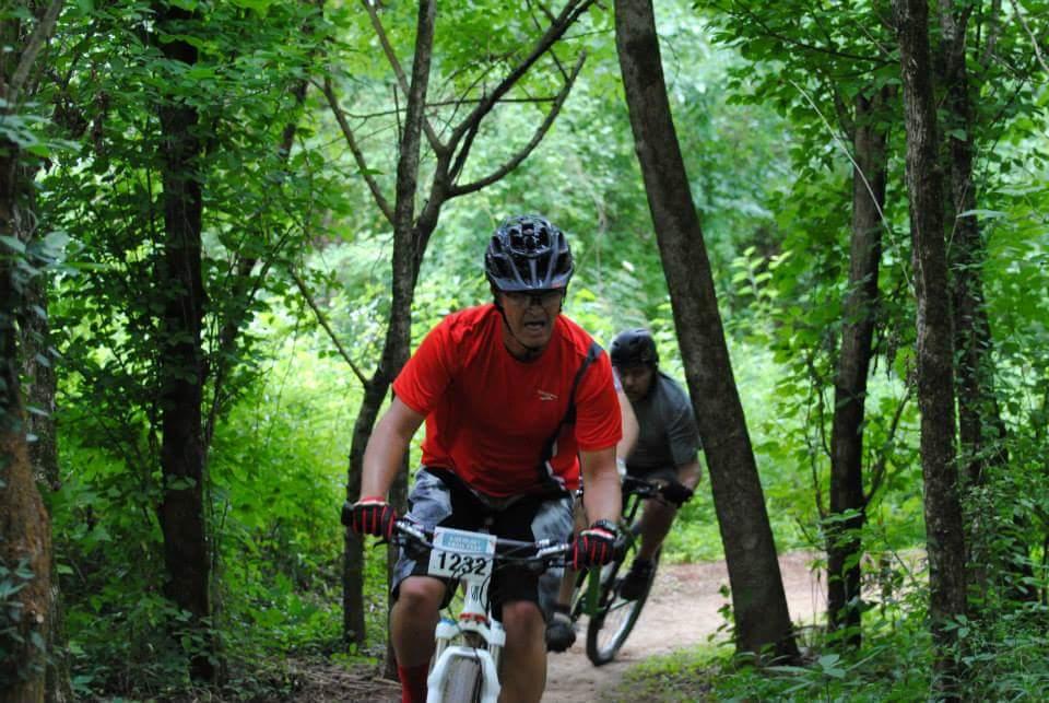 Two mountain bikers navigate a wooded trail. The rider in the foreground, wearing a red shirt and a black helmet, displays a focused expression as he pedals intensely. The second rider, dressed in gray, follows closely behind among the lush greenery and trees. The scene captures the excitement of outdoor cycling in a natural setting. Low Hollow mountain bike trail.