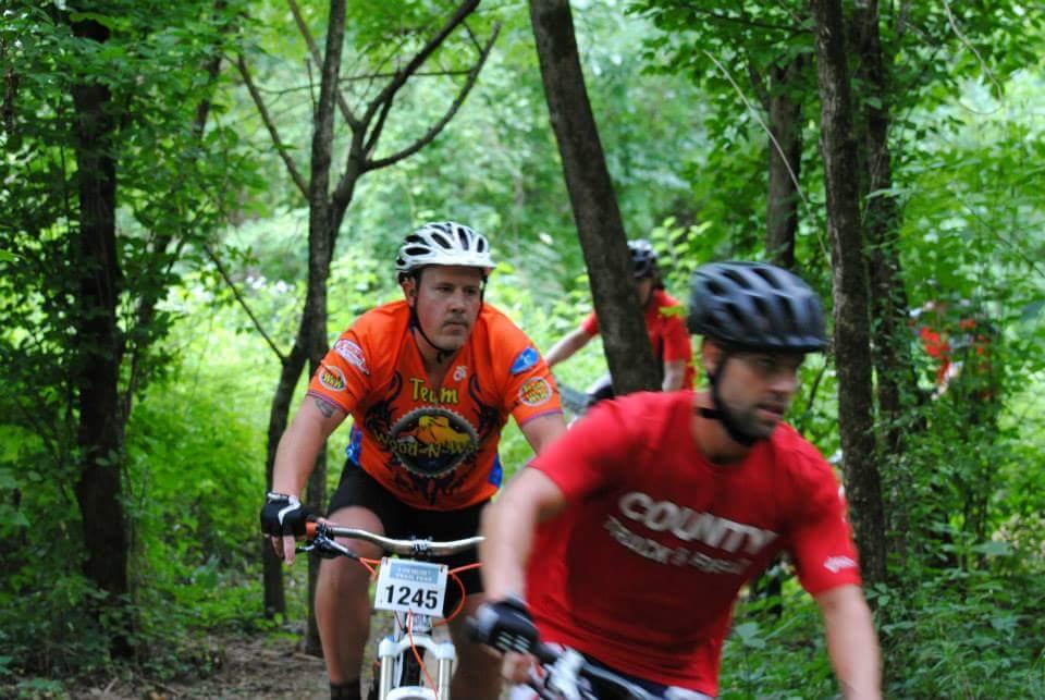 Two mountain bikers ride through a wooded trail. The biker in the foreground wears a red shirt with white text and a black helmet, while the biker behind him wears an orange jersey with various logos. Lush green foliage surrounds the path, creating a natural and vibrant outdoor setting. Low Hollow mountain bike trail.