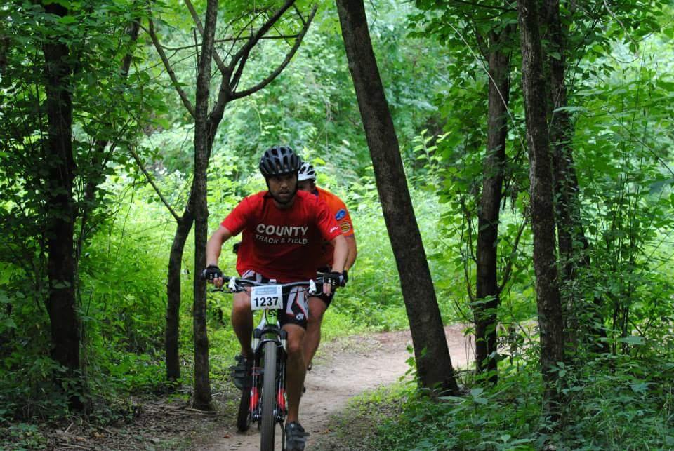 A cyclist wearing a red t-shirt with "COUNTY TRACK & FIELD" printed on it rides on a dirt trail surrounded by lush green foliage and trees. The scene captures the action of mountain biking in a natural outdoor setting. Low Hollow mountain bike trail.