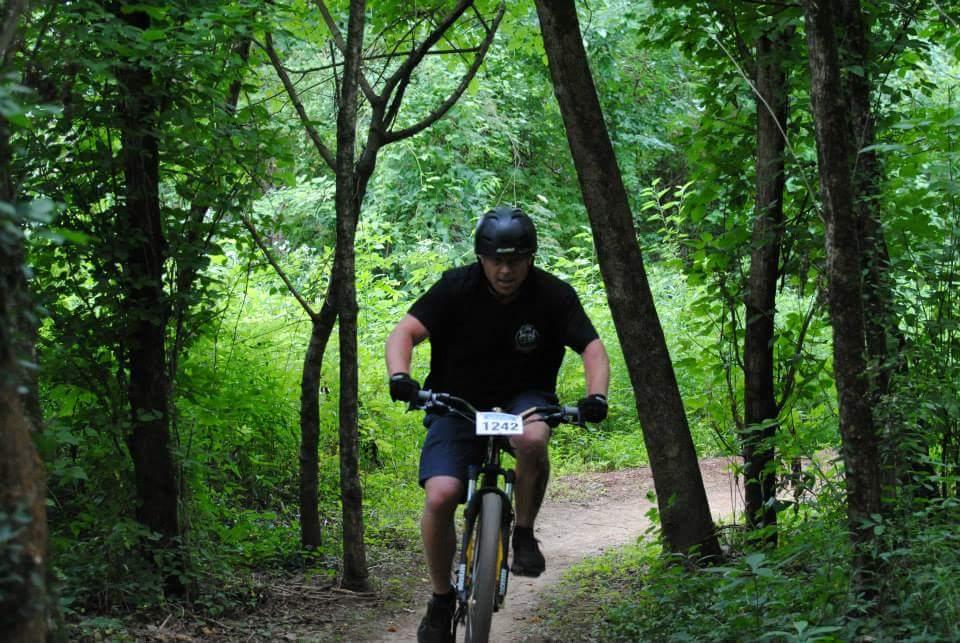 A mountain biker wearing a helmet and a black shirt rides through a lush, green forest trail surrounded by trees and dense foliage. The cyclist appears focused and energetic as he navigates the winding path. Low Hollow mountain bike trail.