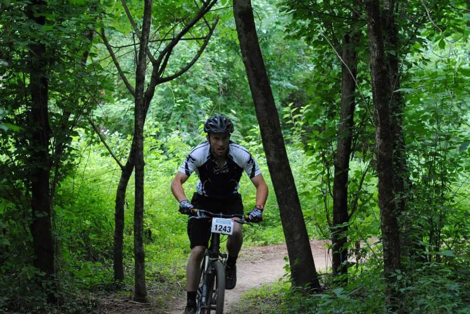 A mountain biker wearing a black and white jersey and a helmet rides along a dirt path framed by trees and lush green foliage. The cyclist is focused and engaged, with a number tag displayed on their jersey. Low Hollow mountain bike trail.