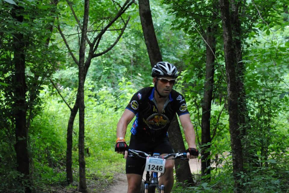A mountain biker riding along a narrow trail through a lush, green forest, wearing a black cycling jersey with colorful designs and a helmet. The cyclist is focused, navigating past trees and thick vegetation. A race number is visible on the bike. Low Hollow mountain bike trail.