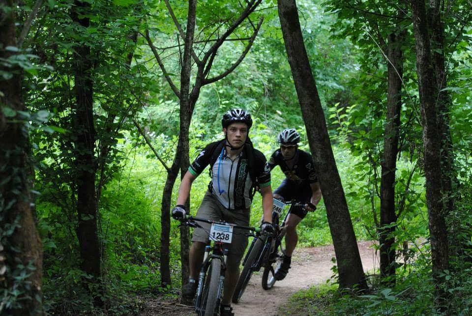 Two mountain bikers riding on a dirt trail surrounded by lush greenery and trees. One cyclist, wearing a black and blue jersey with the number 1238, is in the foreground, while another biker, dressed in black with the number 246, follows behind. The scene captures an active outdoor moment in a natural setting. Low Hollow mountain bike trail.