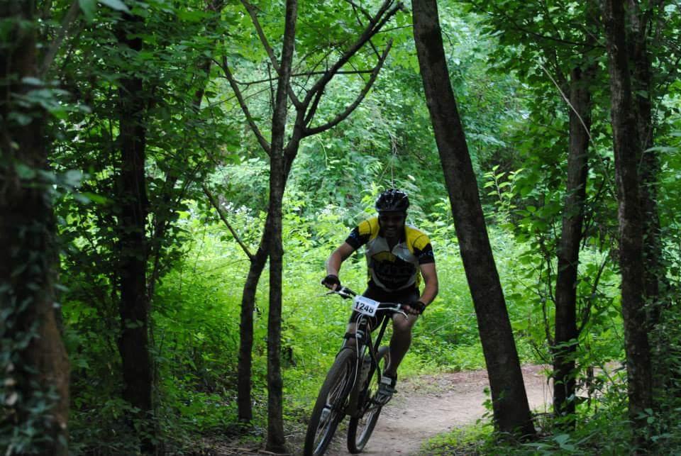 A mountain biker wearing a yellow and black jersey races along a dirt trail in a dense forest, surrounded by lush green foliage and trees. The rider is focused, navigating the winding path on a mountain bike, with a race number visible on their jersey. Low Hollow mountain bike trail.