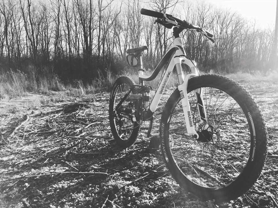 A mountain bike positioned on a dirt trail surrounded by tall, bare trees. The image is presented in black and white, highlighting the bike's details and the textured ground. Sunlight casts gentle shadows, creating a serene outdoor atmosphere. Veterans Park mountain bike trail.