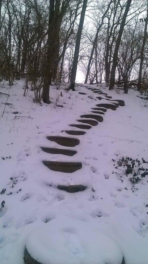 A winding stone pathway leading up a snowy hill, surrounded by bare trees, with fresh snow covering the ground and the path. The scene is calm and quiet, suggesting a serene winter landscape. Veterans Park mountain bike trail.