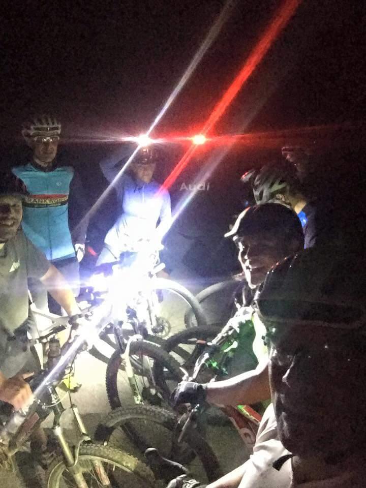 A group of mountain bikers gathered at night, equipped with headlamps and bike lights. The scene is illuminated by bright lights, showcasing the cyclists and their bikes as they pose for a photo. The atmosphere is casual and friendly, highlighting a shared passion for night biking. Veterans Park mountain bike trail.