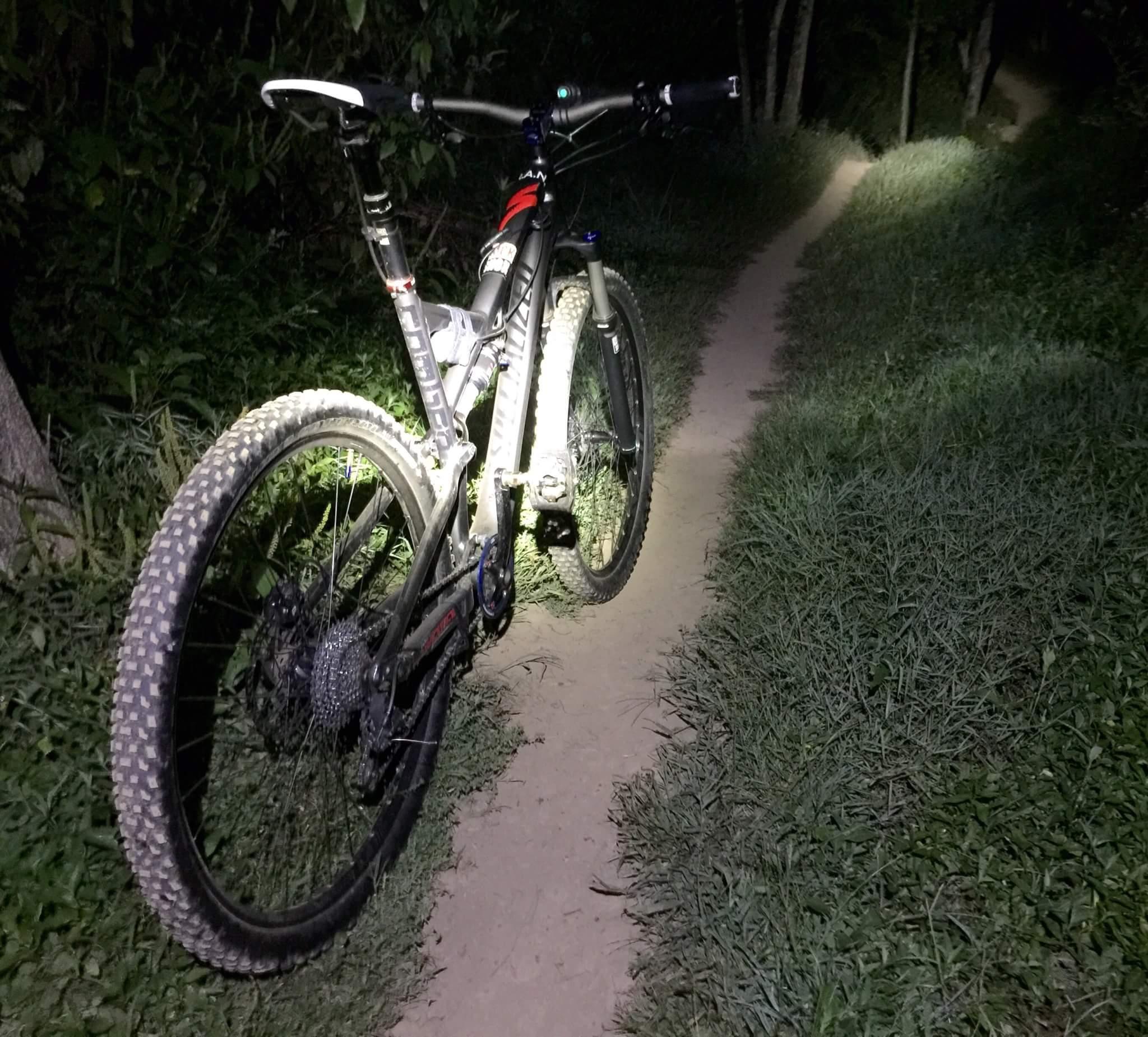 A mountain bike stands on a narrow dirt trail surrounded by tall grass and foliage, illuminated by a bright light. The scene is set at night, highlighting the bike's details with the path winding into the darkness ahead. Veterans Park mountain bike trail.
