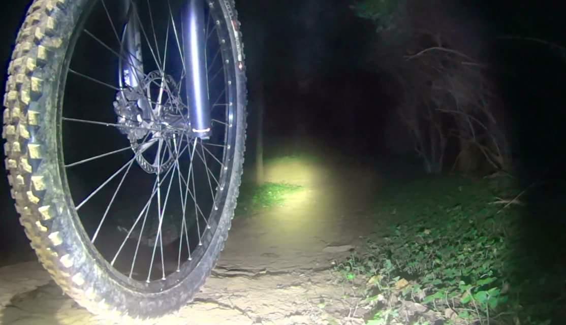 Close-up of a mountain bike tire on a dirt path at night, illuminated by a front light. Surrounding vegetation is faintly visible in the dark background. Veterans Park mountain bike trail.