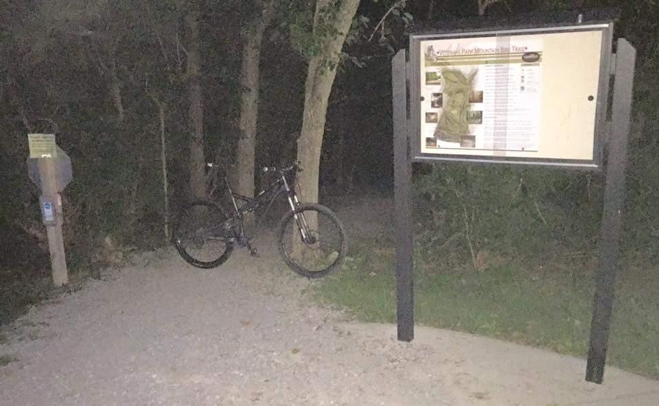A mountain bike rests against a trail sign in a dimly lit wooded area, with trees lining the path. The sign displays information about the trail, while a parking sign is visible nearby. The scene captures a quiet entrance to a biking or hiking trail at night. Veterans Park mountain bike trail.