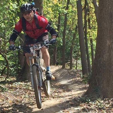 A mountain biker in a red jersey and black shorts rides down a wooded trail, showcasing an exciting moment with both wheels off the ground. The surrounding trees display vibrant autumn foliage. A race number is visible on the biker's jersey. Devou Park mountain bike trail.