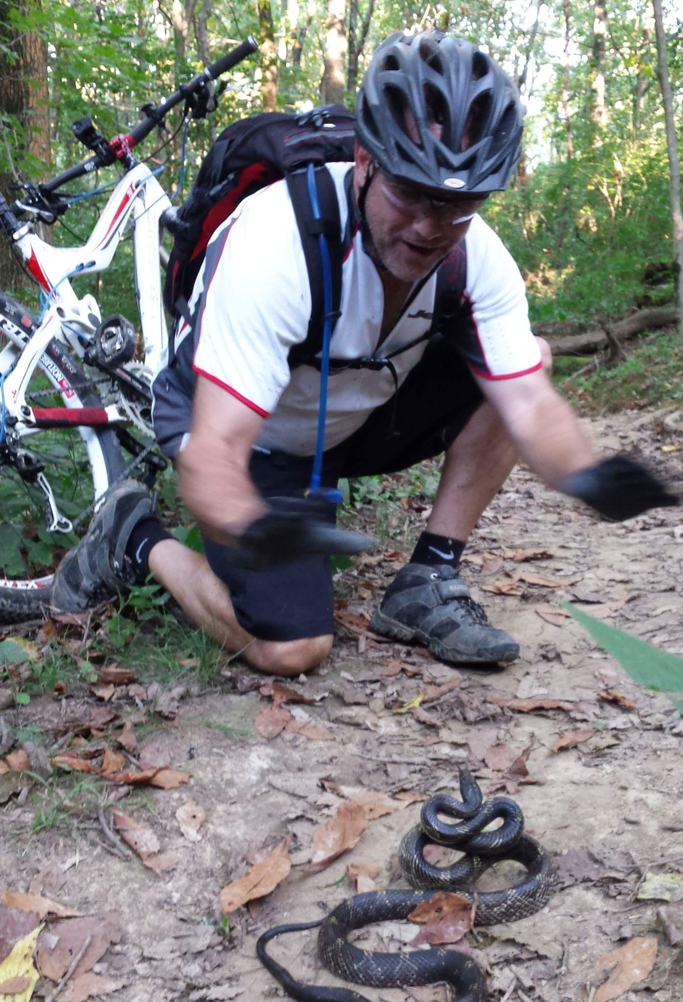 A mountain biker kneels on a dirt trail in a wooded area, appearing to interact with a snake on the ground. The biker is wearing a helmet, sunglasses, and a white cycling shirt, and has a backpack. A bicycle is resting nearby among the trees and fallen leaves. Devou Park mountain bike trail.
